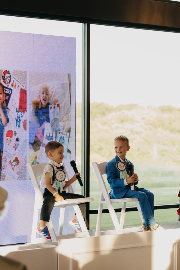 Two boys sit on a stage, one in a blue suit holding a microphone, beside a screen of children’s drawings