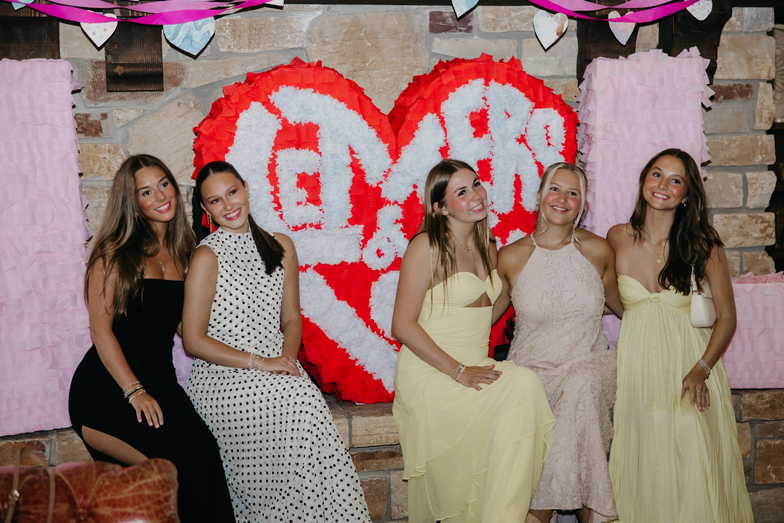 Five young women in formal dresses sit in front of a red and white paper heart sign at a Letters of Love gala, photographed by a nonprofit event photographer in Minneapolis