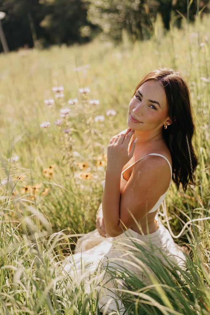 Senior with dark hair pulled back, in a white dress, touching her collarbone in tall wildflowers
