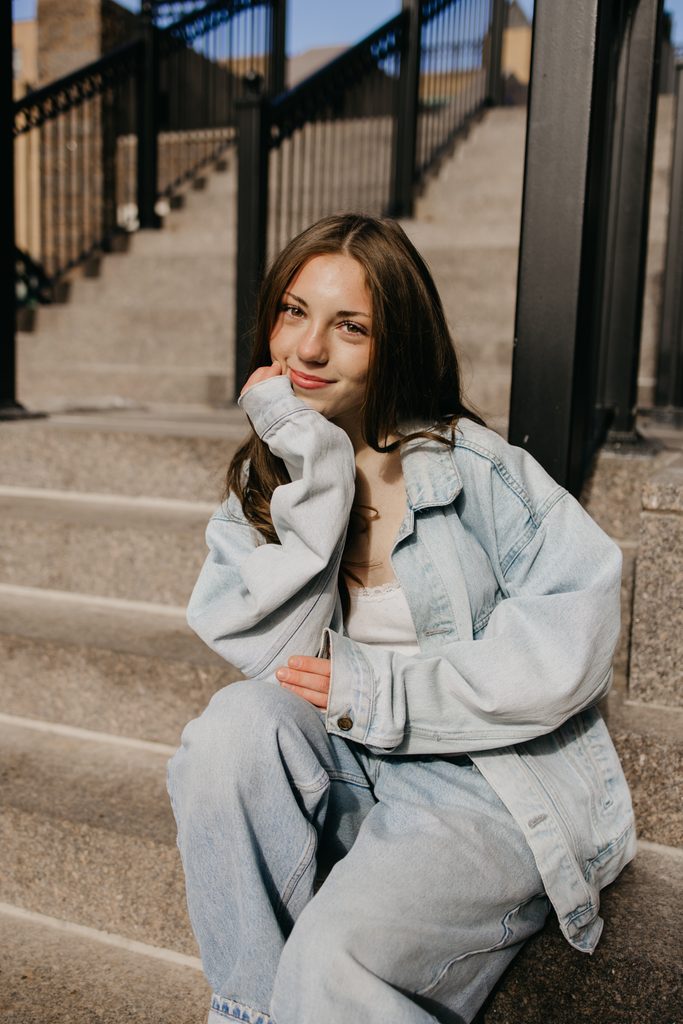 Senior in an oversized denim jacket and light jeans sitting on stone steps, chin resting on her hand