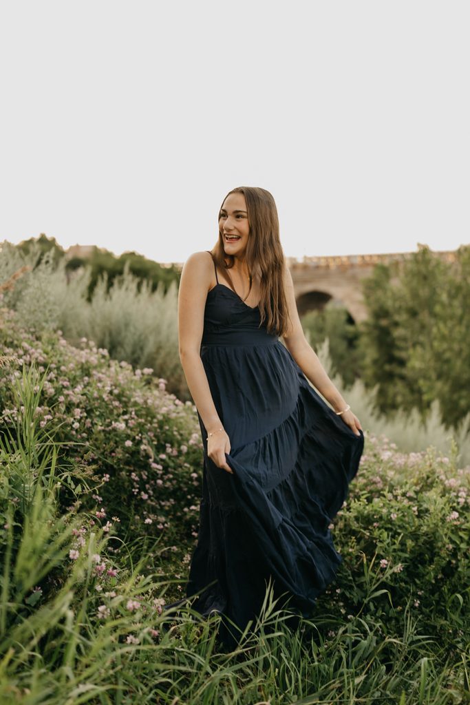 Senior in a long navy dress standing in tall grass with a stone arch bridge behind her