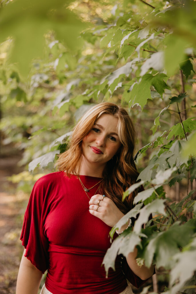 Senior girl in a red flutter-sleeve top smiling beside green leaves during Minneapolis senior pictures
