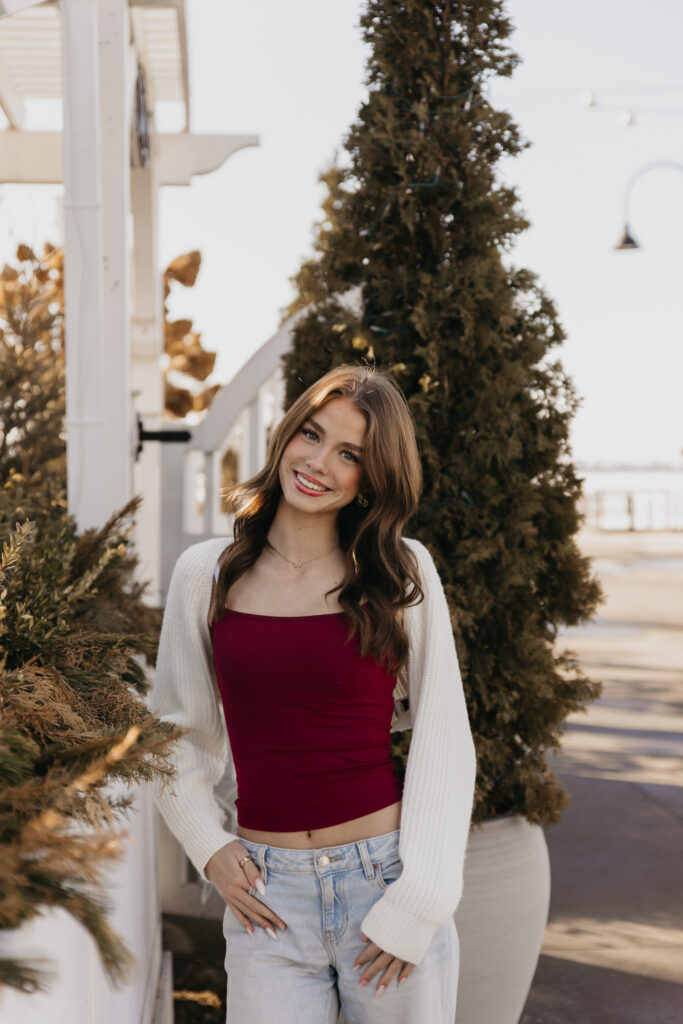 Senior in a burgundy top and cream cardigan posing beside an evergreen near a lakeside pier