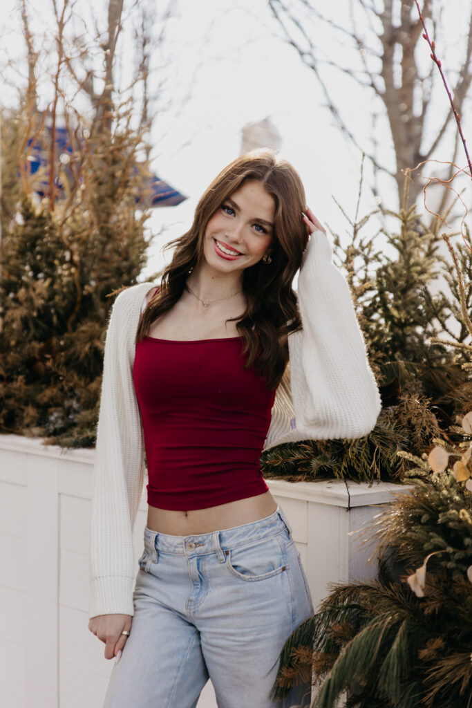 Brunette senior in a burgundy crop top and white cardigan smiling beside winter planters