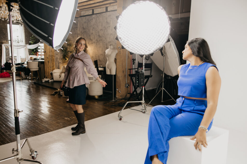 Photographer laughing behind her camera while a client in blue sits on a white studio block