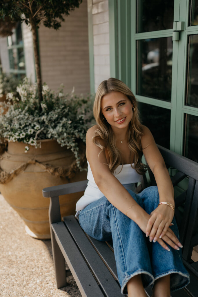 Senior girl in a white tank and wide-leg jeans sitting on a bench beside potted plants