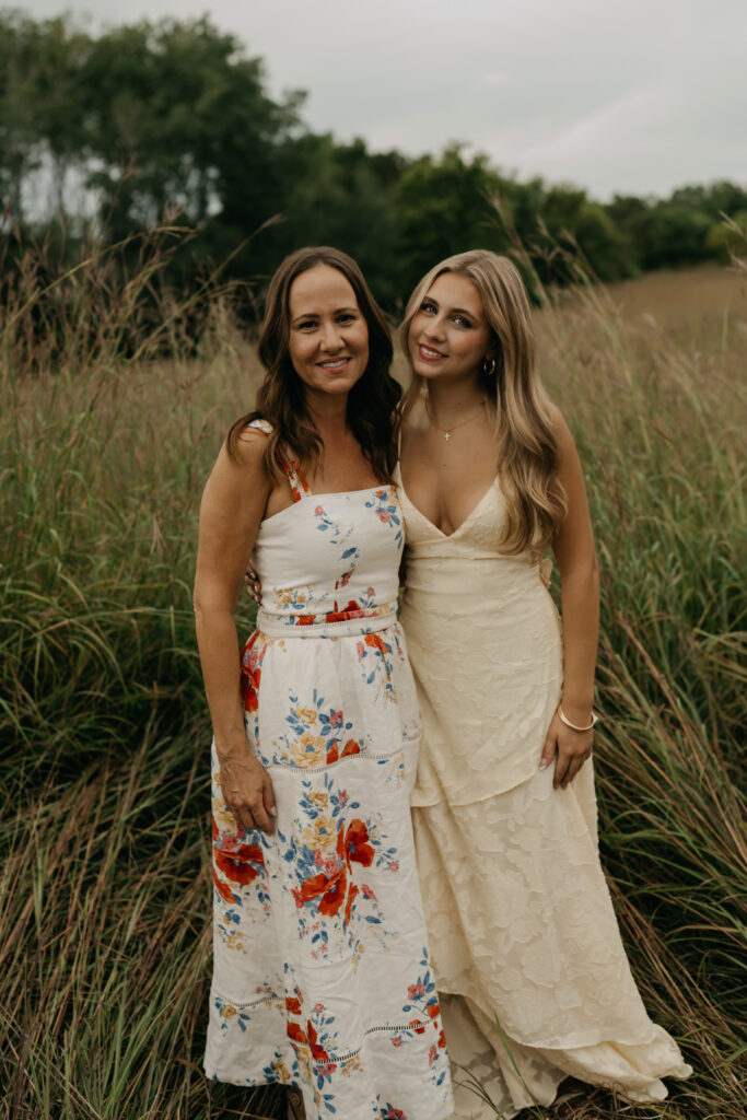 Mom and senior daughter in floral and cream dresses standing together in tall prairie grass