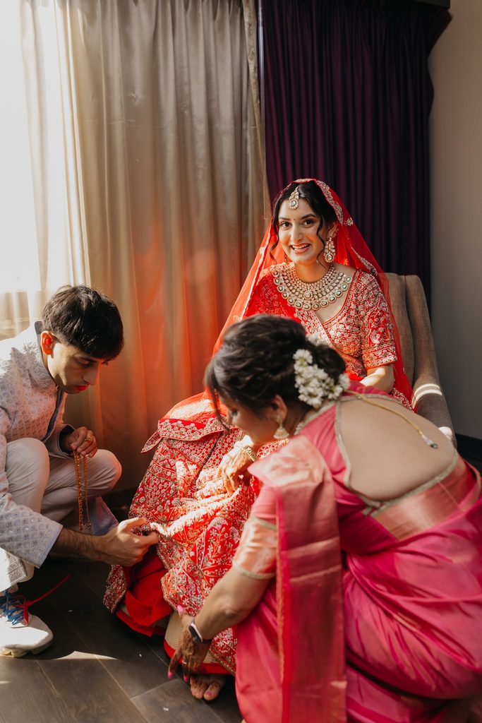 Bride in a red lehenga and veil smiles while family members help her prepare for the ceremony