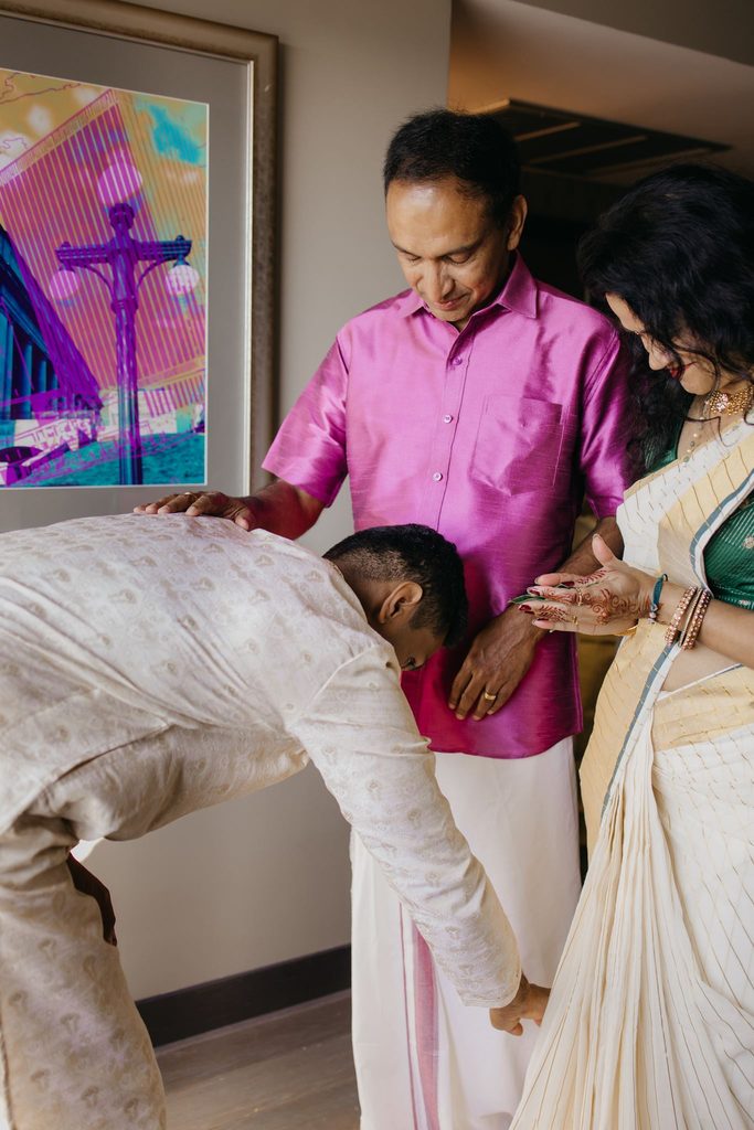 Groom bends to touch the feet of an older man in a pink shirt as a woman in a cream sari watches