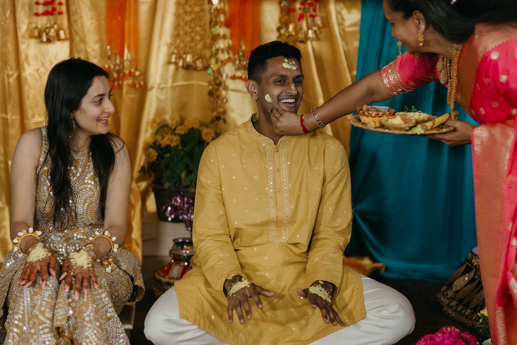 Groom in a yellow kurta with turmeric on his face as a relative holds a plate and the bride smiles beside him