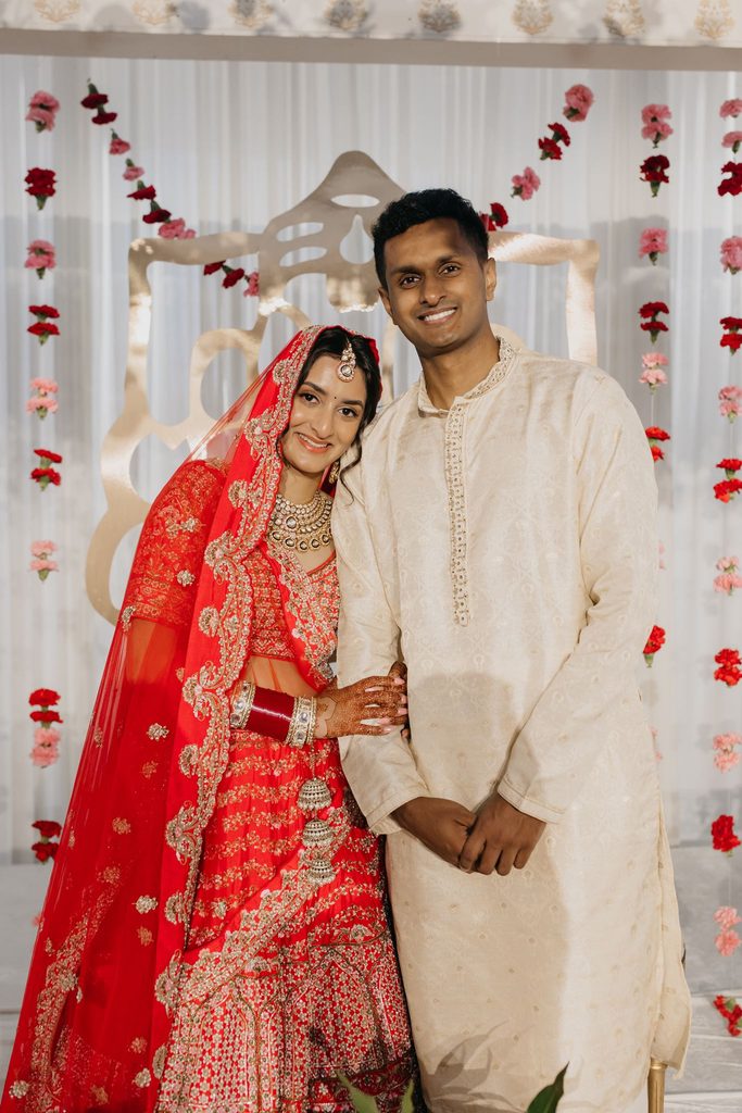 Bride in a red lehenga and veil poses with the groom in a cream kurta before a red and white floral backdrop