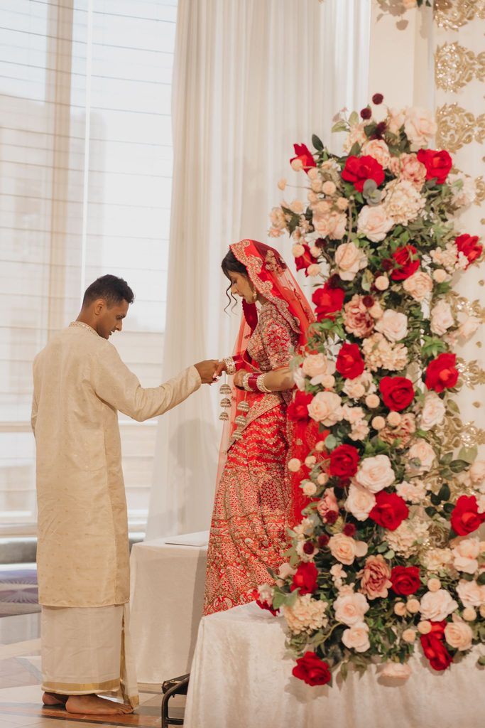Groom in a cream kurta and bride in a red veil stand beside a tall red and white floral arrangement