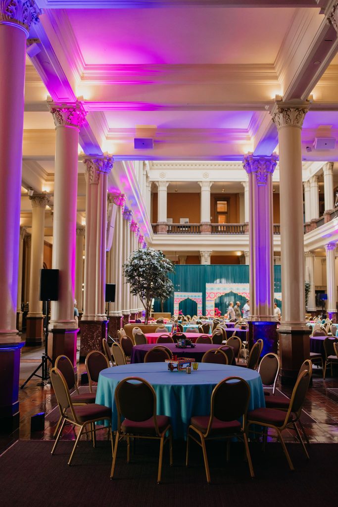 Ballroom with tall columns, purple uplighting, and round tables with teal linens set for an Indian wedding reception
