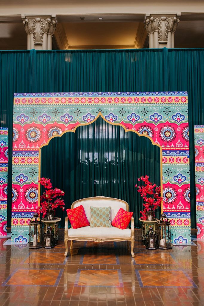 Teal curtain with a colorful arched backdrop, white sofa, and red florals as a sangeet photo spot