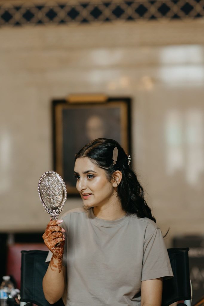 Bride in a gray t-shirt holds an ornate hand mirror with mehndi on her palm while her hair is styled