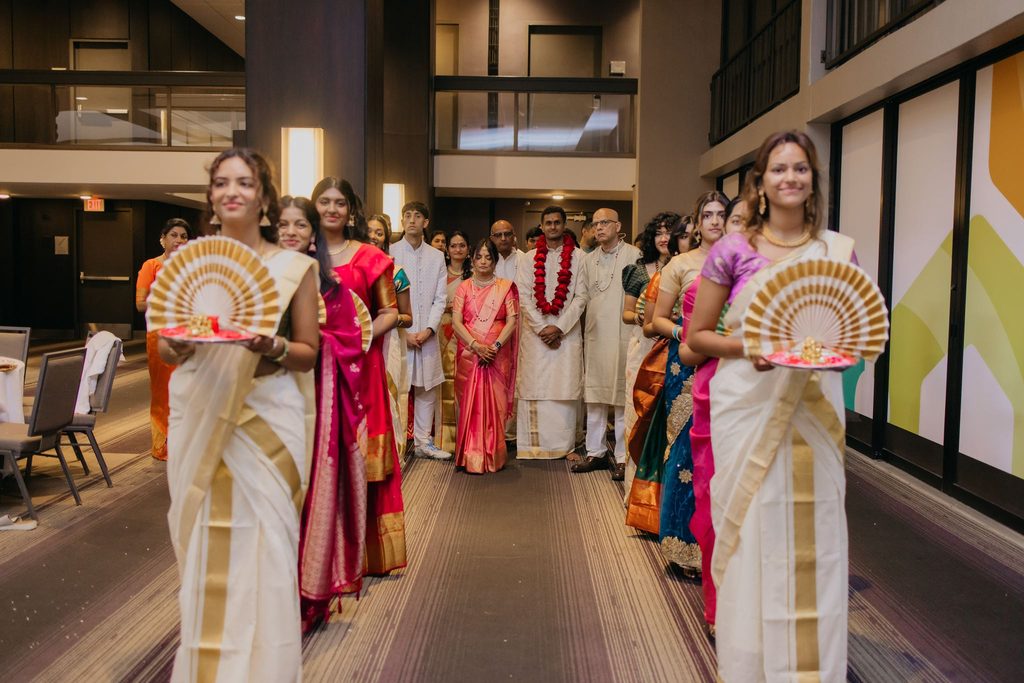 Women in white and gold saris carry decorative fans as they lead a procession of family members