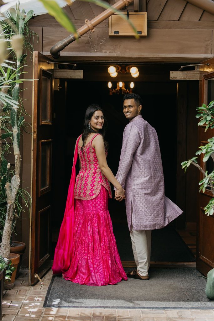 Couple in a pink lehenga and lavender sherwani hold hands and look back from a wooden doorway