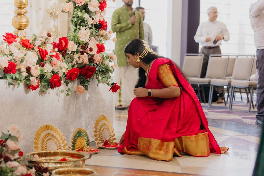 Woman in a red and gold silk sari kneels beside a floral display with brass plates and decorative fans