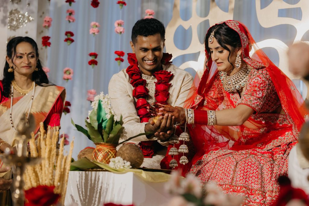 Groom and bride join hands over ritual items during their Indian wedding ceremony as a family member looks on