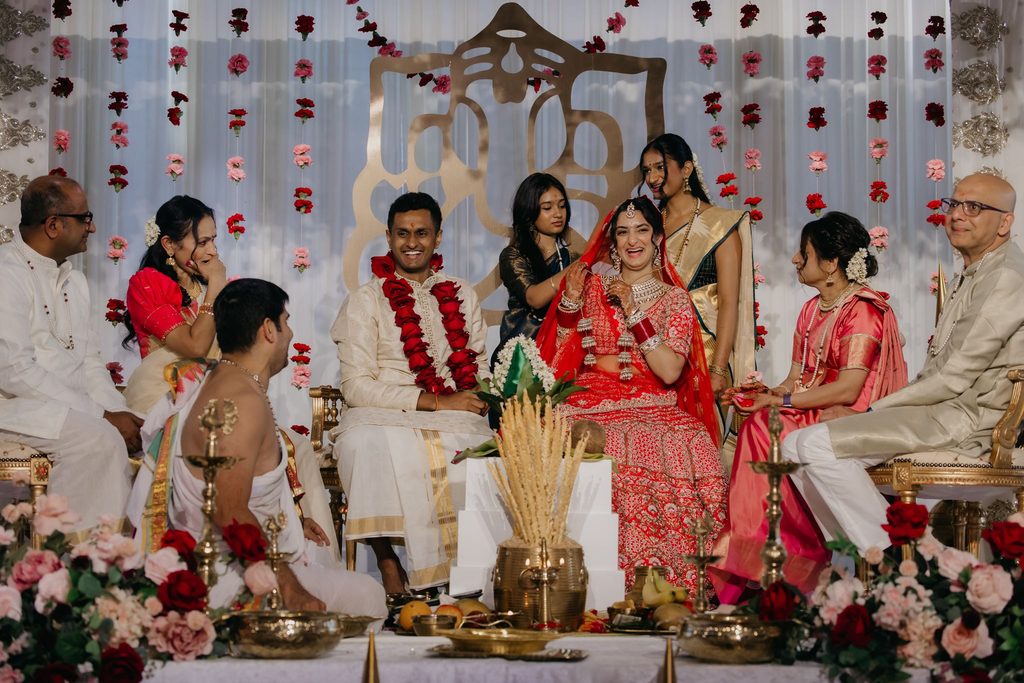 Bride in red and groom in a flower garland seated at an Indian wedding ceremony with family and a priest beside them