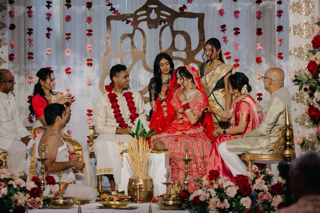 Wide view of the Hindu wedding ceremony with the couple seated at the mandap, red flowers and white drapes behind them