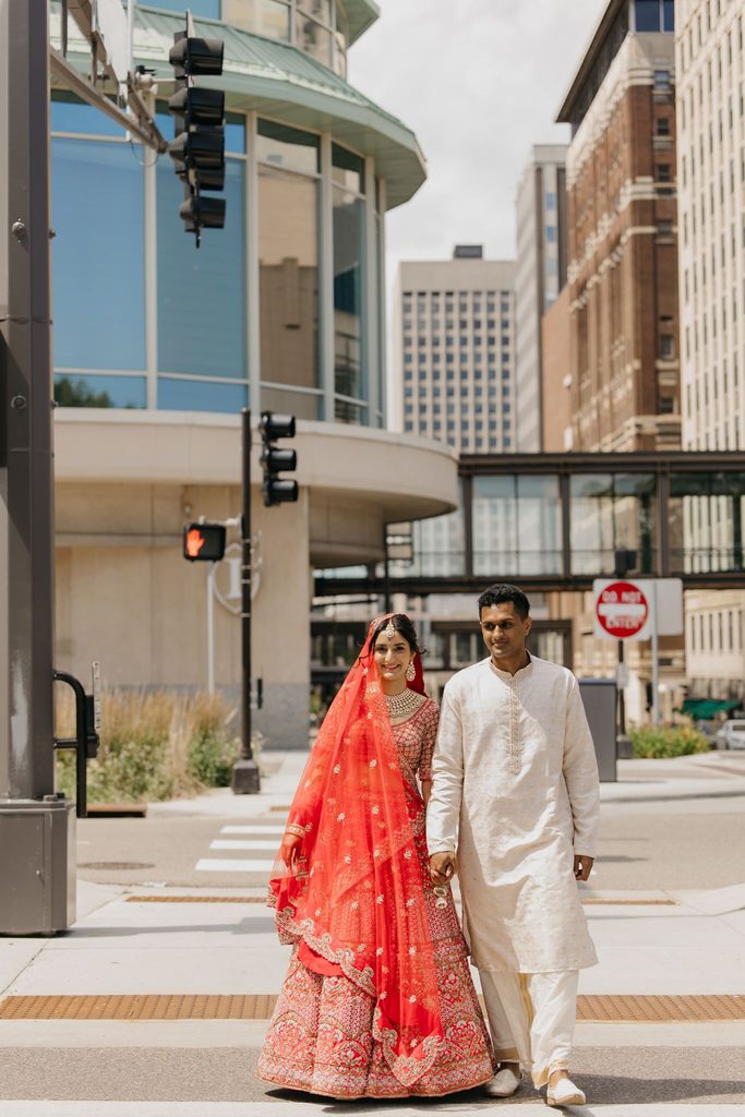 Bride in a bright red veil and groom in a white kurta wait at a downtown Minneapolis street corner