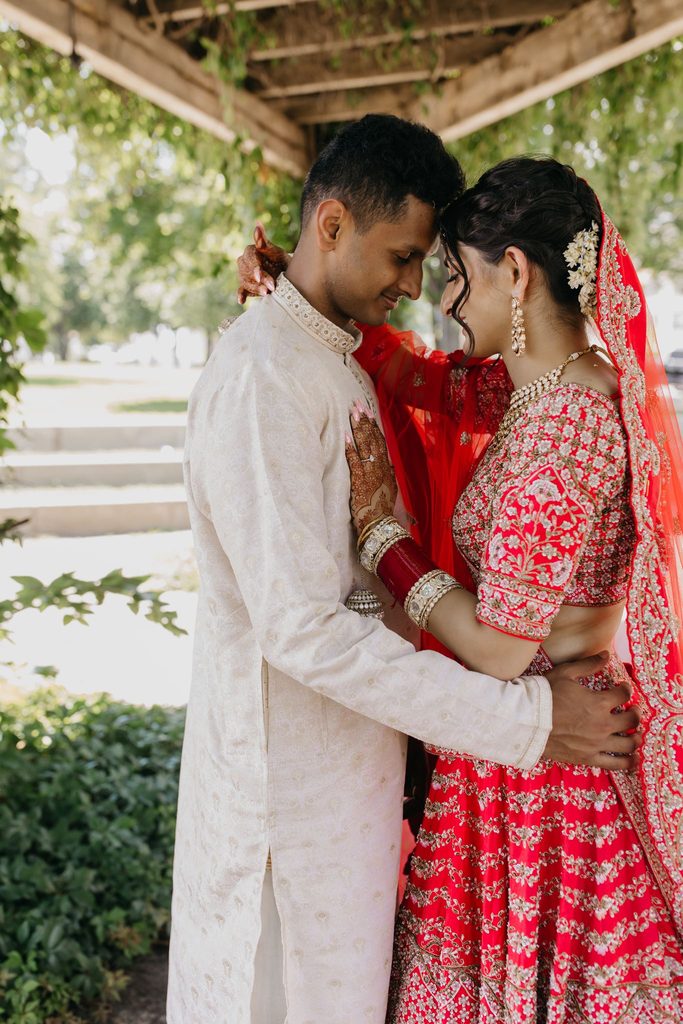 Bride in red and groom in cream embrace under a wooden pergola with their foreheads touching