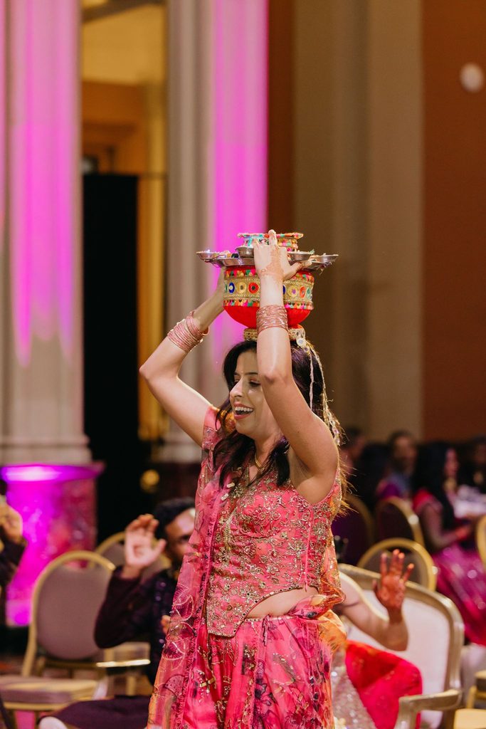 Woman in a pink lehenga dances with a decorated pot balanced on her head during a sangeet performance