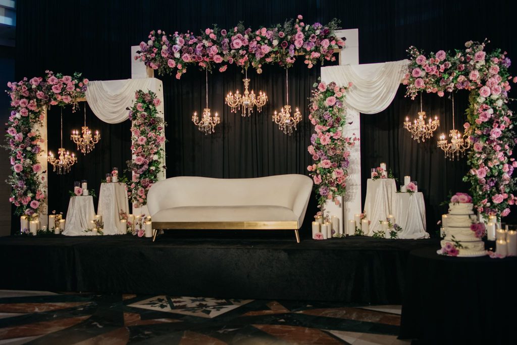 Reception stage with a white sofa, pink and purple florals, and hanging chandeliers against a black backdrop