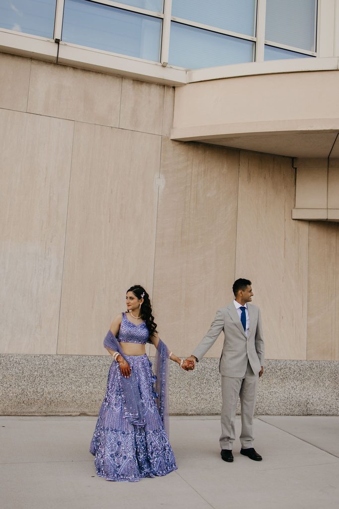 Couple holds hands in full-length view along a stone wall, the bride in lavender and the groom in a gray suit