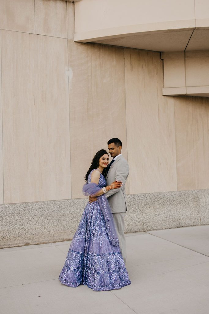 Bride in a lavender sequined lehenga and groom in a gray suit pose against a tan stone wall