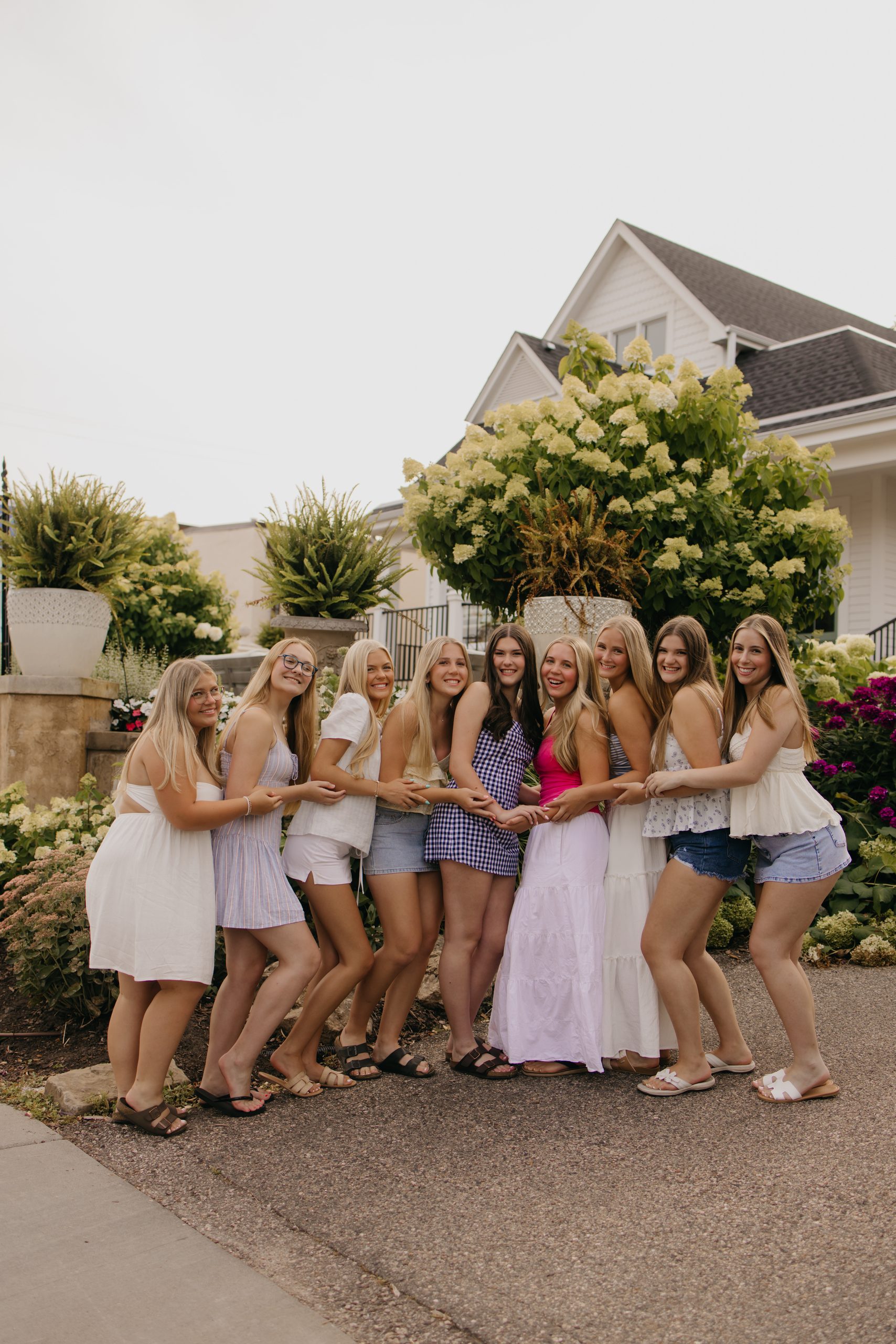 Nine high school seniors in summer outfits link arms and smile in front of hydrangea bushes at a senior rep team group shoot