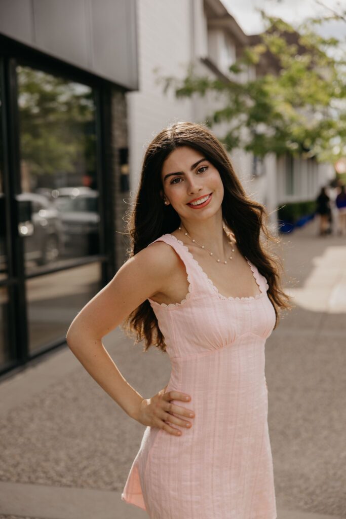 Senior in a pink sleeveless dress posing with a hand on her hip on a city sidewalk