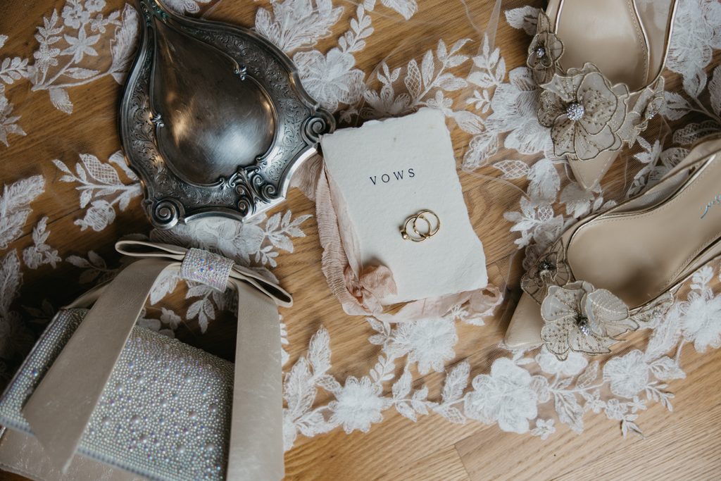 Wedding rings on a vows booklet beside floral heels, an ornate mirror, and a beaded clutch