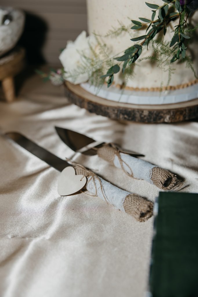 White wedding cake on a wood slice stand beside a heart-tagged cake server and knife
