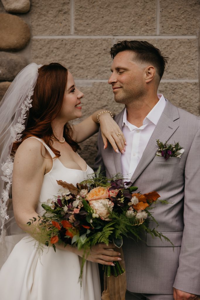 Seth and Stephanie smiling at each other with a fall bouquet in front of a stone wall