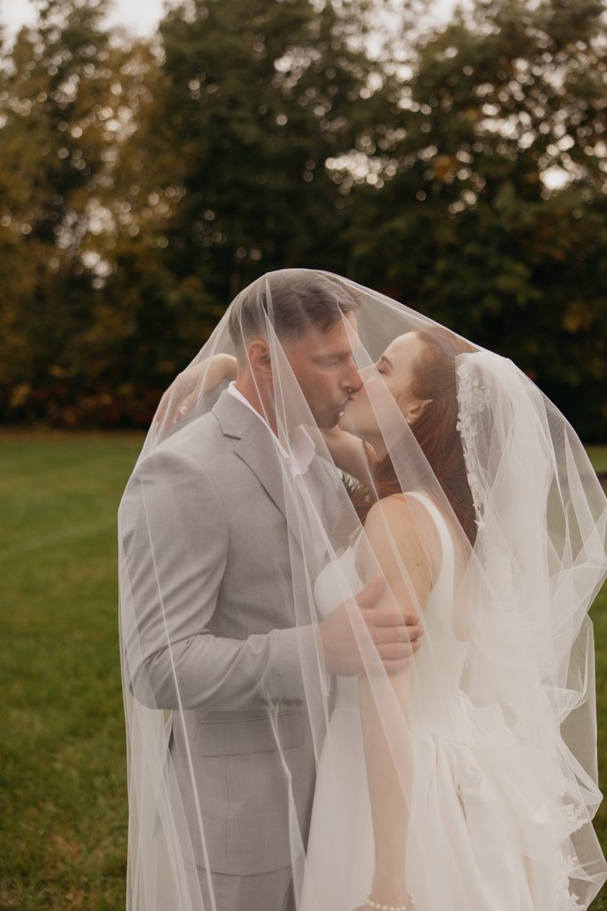 Stephanie and Seth kissing under her sheer lace-edged veil on a green lawn