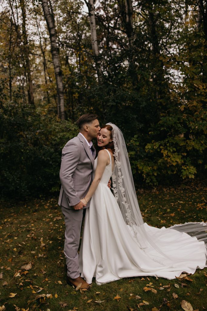 Seth kissing Stephanie’s cheek in a fall woodland clearing, her gown trailing on the grass