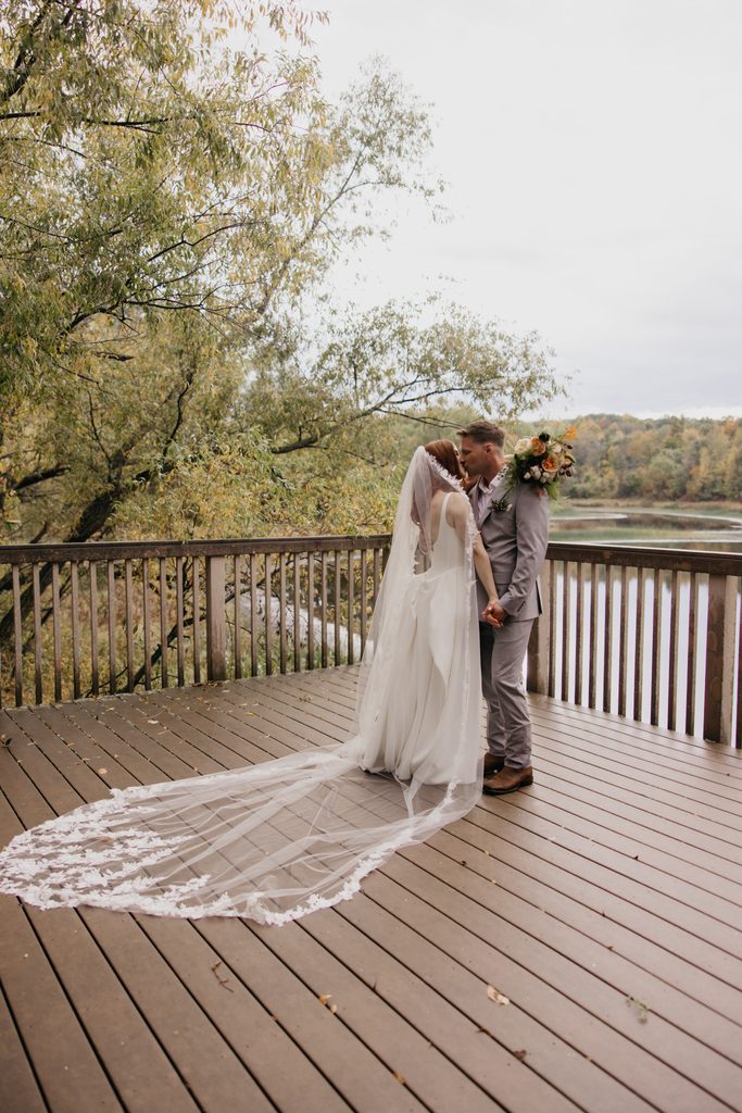 Seth and Stephanie kissing on the deck, her lace veil spread out behind them above the water