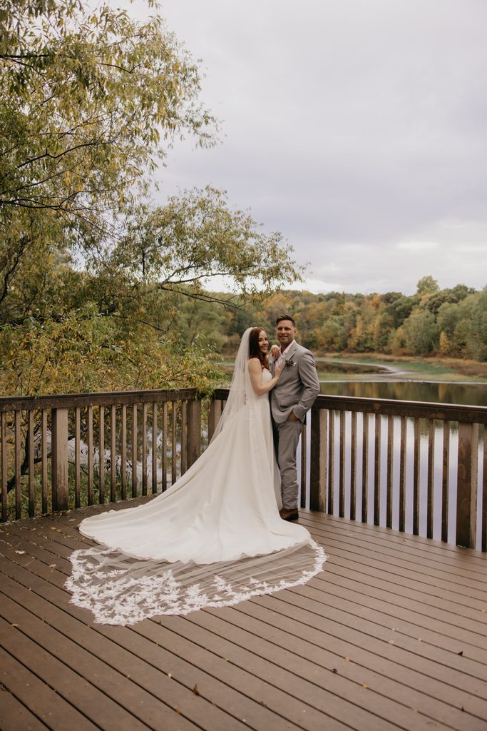 Stephanie and Seth on a deck overlooking the river and autumn trees, her lace-trimmed veil across the boards
