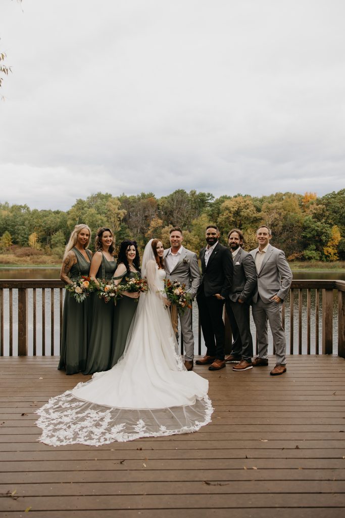 Stephanie and Seth with their wedding party on a lakeside deck at Hidden Lake Park, her long veil spread behind