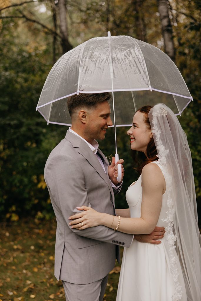 Stephanie and Seth embracing under a clear dome umbrella in the fall woods