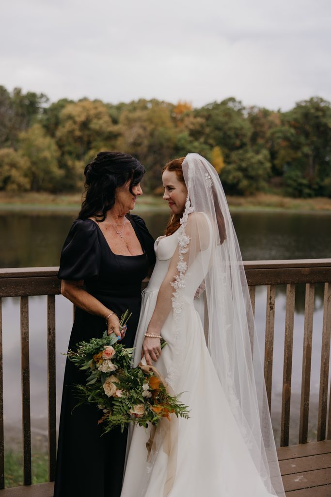Stephanie in her veil smiling at a woman in a black dress on the deck by the lake