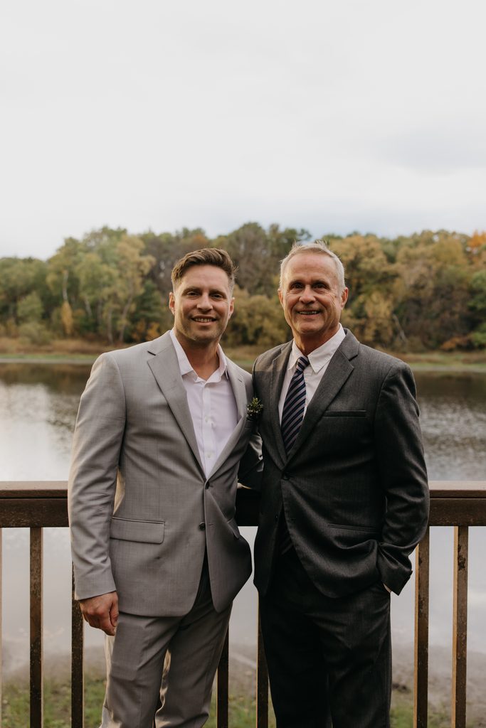 Seth standing beside an older man in a dark suit on a deck above the lake