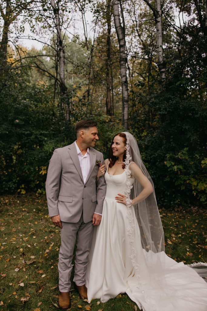 Stephanie and Seth standing side by side among birch trees, smiling at each other