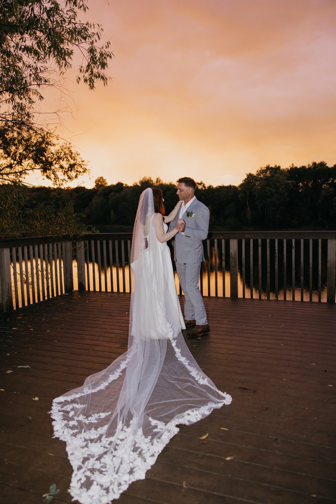 Bride and groom on a deck at sunset at Hidden Lake Park, her lace veil spread behind
