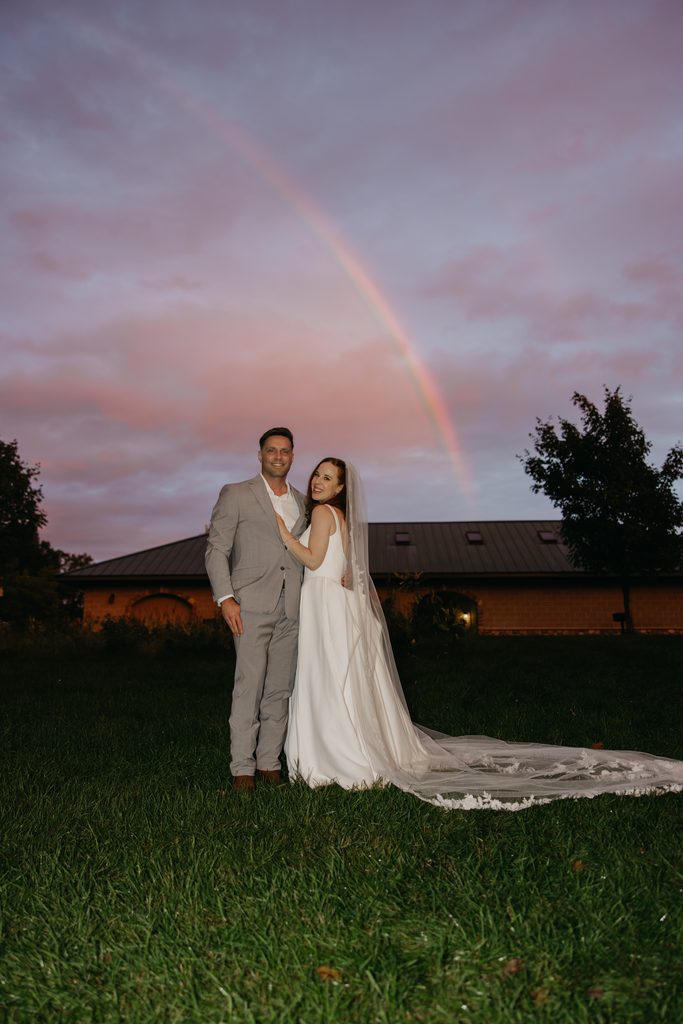 Stephanie and Seth on a lawn under a pink dusk sky with a rainbow