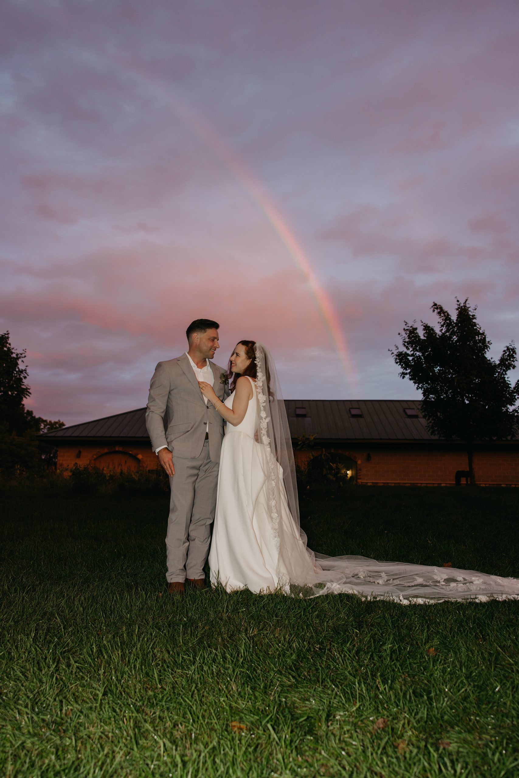Bride and groom embrace on a lawn under a pink dusk sky with a rainbow