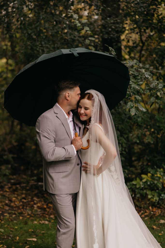 Stephanie smiling as Seth kisses her under a dark umbrella in the woods