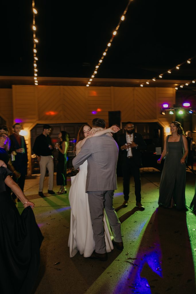 Stephanie and Seth embrace as guests dance under string lights and colored party lights